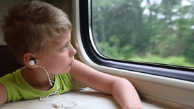 Closeup view video portrait of cute white little kid travelling by train. Boy sits on his seat near window and looks out from window through glass while listening music cheerfully using ear phones. 