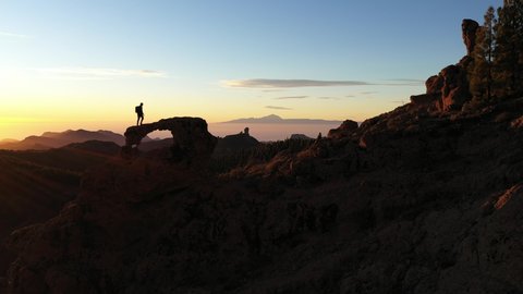 Slow Motion Wide Drone Shot Of Man Walking On Rock Arch Overlooking Mountain Landscape At Sunset