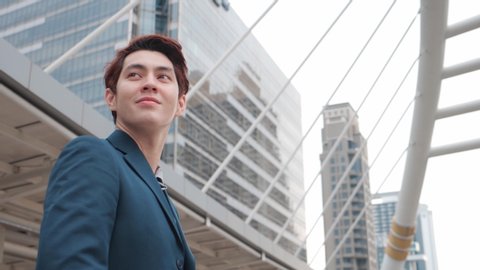 Asian mixed race young businessman in suit looking out while standing near modern Office Building. 4K Slow Motion Corporate Shot with Camera of Moving Around.