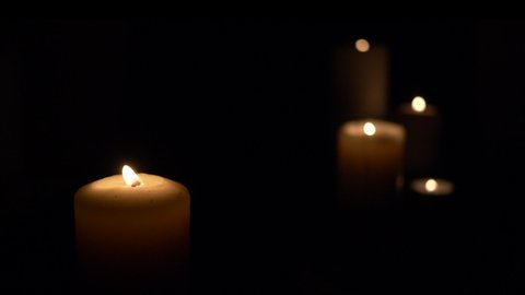 A set of lit white candles flickering in the dark against a black background, with one candle in focus in the foreground.