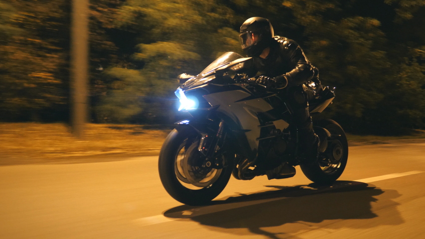 Young man in helmet riding fast on modern black sport motorbike at evening city street. Motorcyclist racing his motorcycle on night empty road. Guy driving bike. Concept of freedom and hobby. Close up