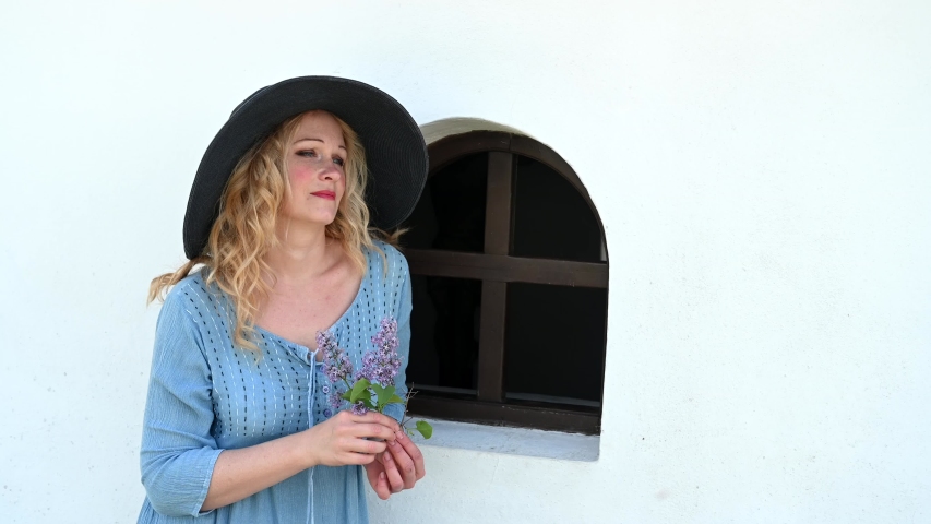 Blond european middle age woman with large hat near window taking off hat in blue dress romantic