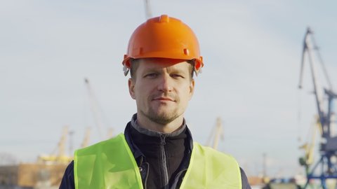 Head and shoulders portrait of middle aged marine engineer in orange helmet looking at camera and greeting you at dockside