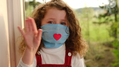 Close up of little girl wearing medical mask with a red heart sitting in windowsill at home vlogger saying hello hi looking at Camera, small kid talking to webcam making online recording having fun