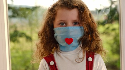 Portrait of cute small kid girl wears facial medical mask with red heart, happy funny little preschool child sitting on windowsill looking at camera. Coronavirus or COVID-19 pandemic disease symptoms