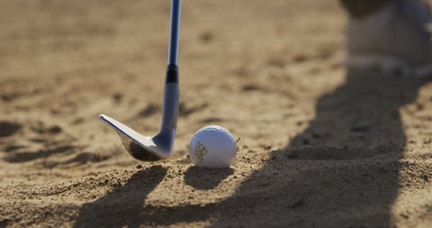 Close up of the club of a golfer on a golf course on a sunny day, swinging and hitting a golf ball in a sand bunker, in slow motion 
