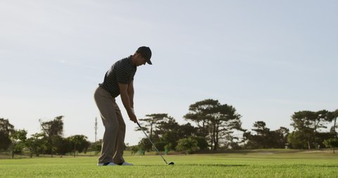 Side view of a Caucasian male golfer on a golf course on a sunny day wearing a cap and golf clothes, swinging a golf club and hitting the ball into the distance, in slow motion 