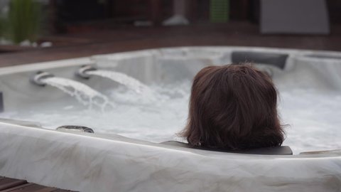Back view of a woman enjoying the jacuzzi. Front view of a young woman enjoying the jacuzzi in a wellness center. Joyful woman resting in a jacuzzi. Woman in spa salon.