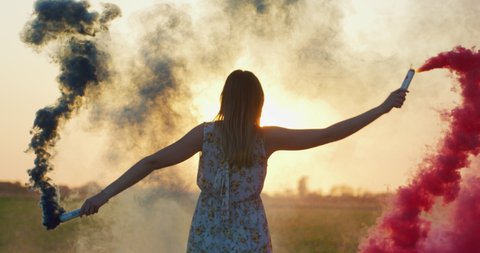 An Young Carefree Happy Woman is Having Fun with Colorful smoke flares in a Countryside on a Sunset.