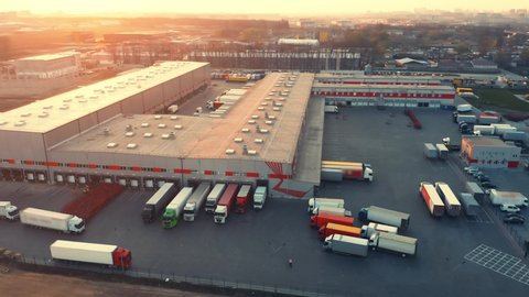 Aerial panoramic view of the big logistics park with warehouse, loading hub with many semi trucks with cargo trailers standing at the ramps for loading/unloading goods at sunset