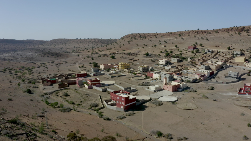 An aerial straif across a small muslim village of colourful adobe houses, surrounded by an arid desert landscape dotted with shrubs, blue sky in the distance.