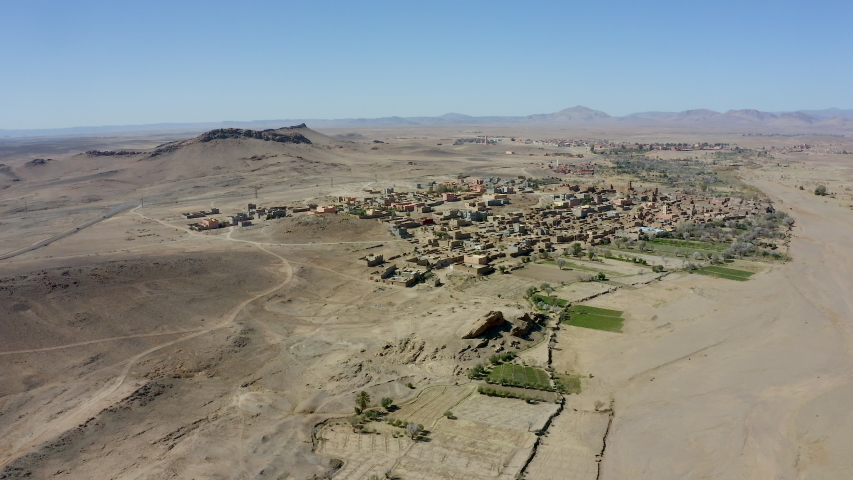 An aerial boom down overlooking a small town in the middle of a vast expanse of barren desert landscape, with a ridge of mountains visible in the distance. Mid morning or afternoon on a bluebird day.