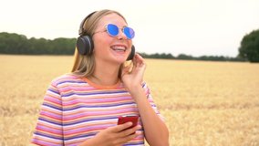 Slow motion video clip of pretty blonde girl teenager young woman wearing a striped t- shirt and blue sunglasses in a field listening to music on her cell phone and wireless headphones 