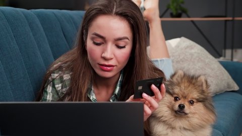 Calm relaxed Woman is lying on Sofa in Livingroom with her Dog while getting Online Shopping with Credit Card. Young Woman is ordering Purchases online. Shopping Online Indoor. Delivery. Casual Life.