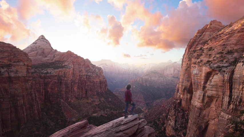 Adventurous Woman at the edge of a cliff is looking at a beautiful landscape view in the Canyon during a vibrant sunset. Taken in Zion National Park, Utah, United States. Parallax Panorama Royalty-Free Stock Footage #1051996741