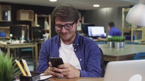 Happy Young Man Uses Smartphone while Sitting at office. Man Browses Through Internet, Watches Videos and Uses Social Networks
