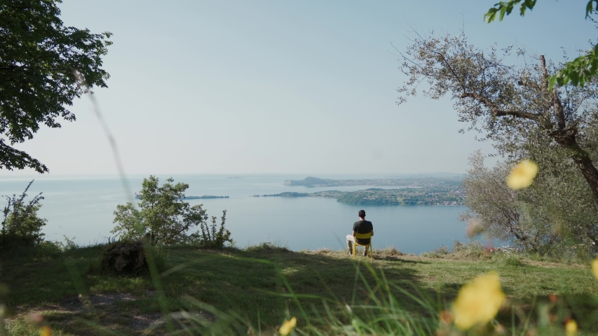 Young man sits on yellow chair on a hill, rests, looks at a beautiful lake landscape on a sunny day. Lake Garda, Italy. Ideal place for rest, relaxation, pondering, daydreaming with a picturesque view