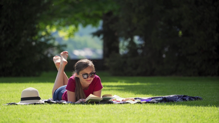 Woman has picnic in green park. Pretty girl in sunglasses enjoy reading book in the garden on sunny day. Woman lies cross-legged on blanket with straw hat Royalty-Free Stock Footage #1052003254