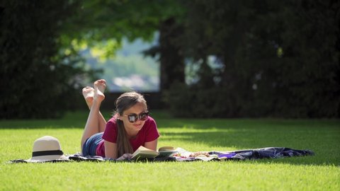 Woman has picnic in green park. Pretty girl in sunglasses enjoy reading book in the garden on sunny day. Woman lies cross-legged on blanket with straw hat