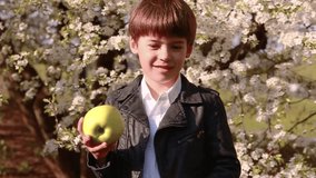A little boy stands in the background of flowering trees in the garden, looks at the camera and twirls a green apple in the rivers. Slow motion video