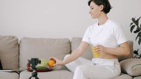 The woman with an orange juice in hands is sitting on a sofa. The woman is filming herself for the vlog and show healthy food.