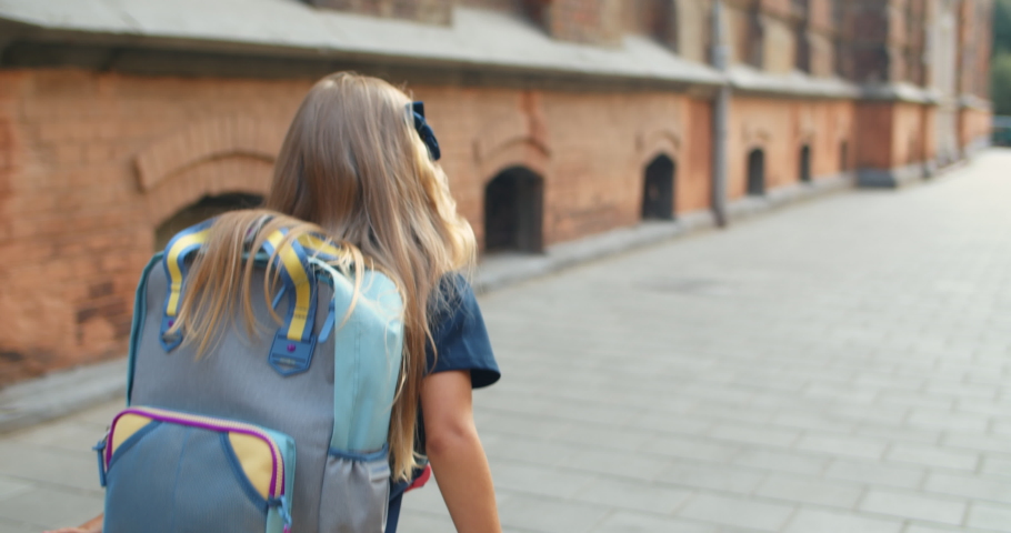 Back view of little blond hair kid with bag on her back jumping happily at street. Schoolgirl going on her way to school. Concept of education. Outdoors