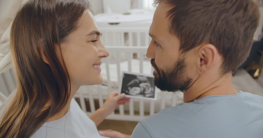 Close up back view of happy young man and pregnant woman hugging and cuddling looking at sonogram image standing in nursery room with cradle. Pregnancy and parenthood concept Royalty-Free Stock Footage #1052018275