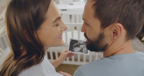 Close up back view of happy young man and pregnant woman hugging and cuddling looking at sonogram image standing in nursery room with cradle. Pregnancy and parenthood concept