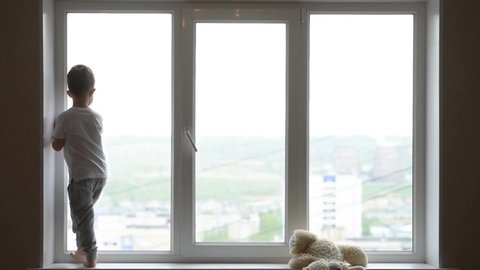 a little boy in a mask sits at home in quarantine and looks out the window in a place with a teddy bear. Prevention of Coronavirus and Covid - 19. Concept