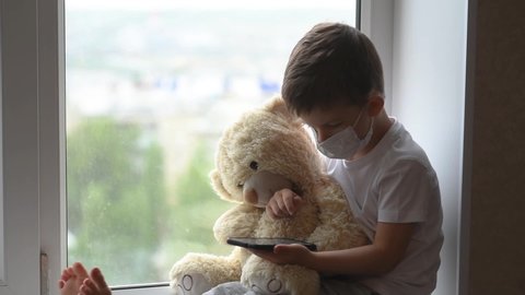 A little boy sits on a window with a teddy bear in quarantine and plays in a mobile phone. Prevention of coronavirus and Covid - 19