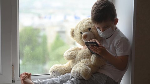 A little boy sits on a window with a teddy bear in quarantine and plays in a mobile phone. Prevention of coronavirus and Covid - 19