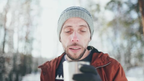 Close-up of a Caucasian man in a gray hat, sweater, brown jacket and gray gloves stands in a winter forest, blows on a hot tea in a mug, takes a sip of tea. Stock Video