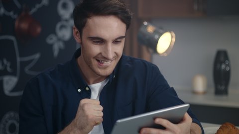 Surprised man getting good news on tablet computer at home office. Cheerful business man reading positive results of project at tablet indoors. Winner male person feeling happy at remote workplace.