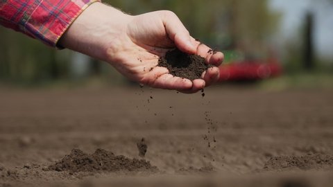 close-up of hand, farmer, agronomist checks quality of soil on farm field. background of working tractor, cultivator. Modern Agriculturally potato cultivation. spring sunny day.