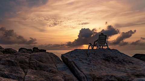 A man sits in a lounger on the rocs by the sea against the background of sunset in summer. 4K time lapse