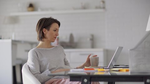 Side view medium shot of beautiful middle aged woman having business video call on laptop computer at home. Businesswoman drinking tea and talking to partner