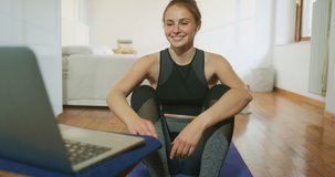 An Young Smiling Blond Hair Woman Wearing Sportswear is Making Fitness Exercises with Training Tutorial Videos on a laptop on a Floor with Mat at Home.