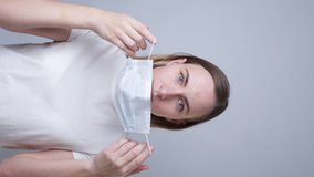 Vertical video. Woman puts a medical mask on her face on a gray background