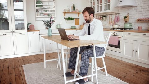Young bearded man working from home with laptop wearing shirt, tie and pajama pants in kitchen, slow motion