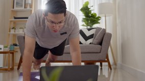 Asian man doing mountain climber plank exercise in living room at home, watching live or video tutorial online via laptop computer. Activity during quarantine and social distance new normal concept.
