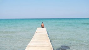Teenager sits on the edge of wooden pier against the backdrop of beautiful azure sea, slow-motion video.