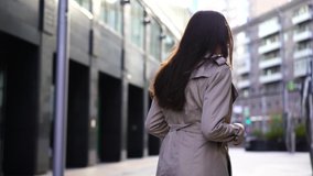 Close-up view from the back of a girl with long brown hair, she is walking, she is wearing a stylish beige coat and jeans.