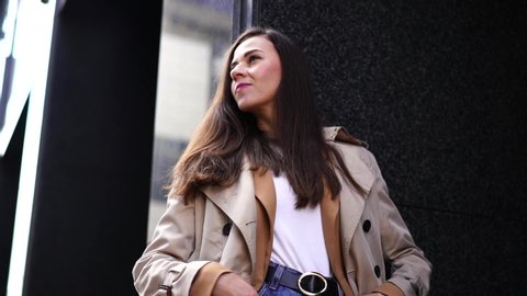 portrait of a woman with long hair, walking around the city against the background of a European office building, standing against the wall and posing, wearing a stylish beige jacket and light coat. : vidéo de stock