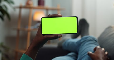 Lviv, Ukraine - 11 February, 2020: Man scrolling smartphone mock up screen while holding it in horizontal landscape mode. Guy watching video blog while lying on sofa. Concept of chroma key and green