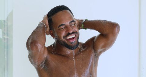 Happy sexy young afro american ethnic man singing taking morning shower. Smiling bearded african muscular hipster guy washing under warm water listening music enjoying hygiene routine in bathroom.