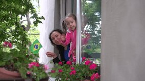 Covid-19. Brazilian hispanic family mother and little girl child on balcony / window are smiling and waving the flag of Brazil. Self-isolation in quarantine, lockdown, stay at home, social distancing - Powered by Shutterstock - Get 15% off with code: PIKWIZARD15
