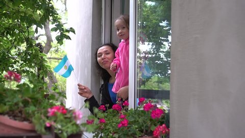 Covid-19 Argentinean hispanic family mother and little girl child on balcony are smiling and waving the flag of Argentina. Self-isolation in quarantine, lockdown, stay at home, social distancing
