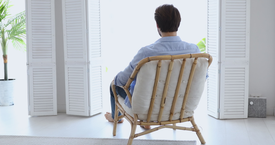 Calm young man relaxing sitting in comfortable wooden chair enjoying lounge lifestyle on peaceful summer day holding hands behind head, breathing fresh air in cozy apartment. Rear back view. Royalty-Free Stock Footage #1052071954