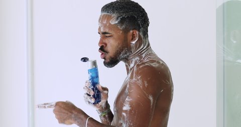 Funny happy young african american man singing in shower gel bottle while taking shower. Smiling handsome ethnic guy listening music having fun enjoying washing hygiene morning routine in bathroom.