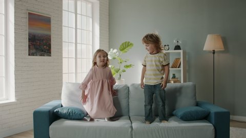 Funny cute caucasian brother and sisiter playing together at home, jumping on couch and happily laughing - family fun, happy childhood concept 4k footage
